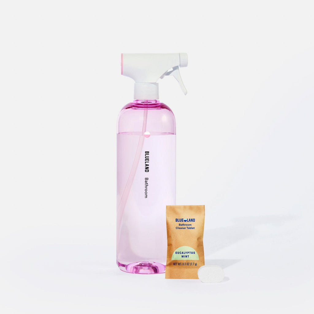 A clear spray bottle labeled Blueland Bathroom with pink liquid inside is next to a packet labeled Blueland Cleaner Tablet Eucalyptus Mint and a pink cleaning tablet on a white background.