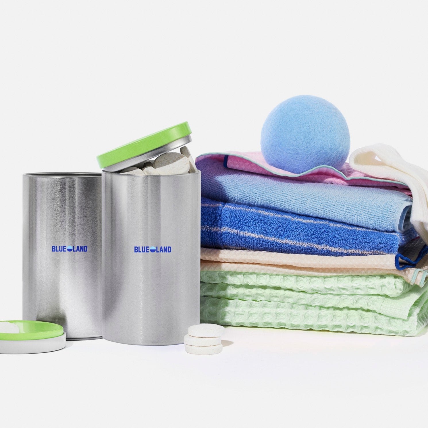 Two pastel-colored Blueland containers labeled “Oxi Laundry Booster” and “Laundry Detergent Tablets” next to laundry tablets, blue dryer balls, and a neatly folded stack of colorful towels.