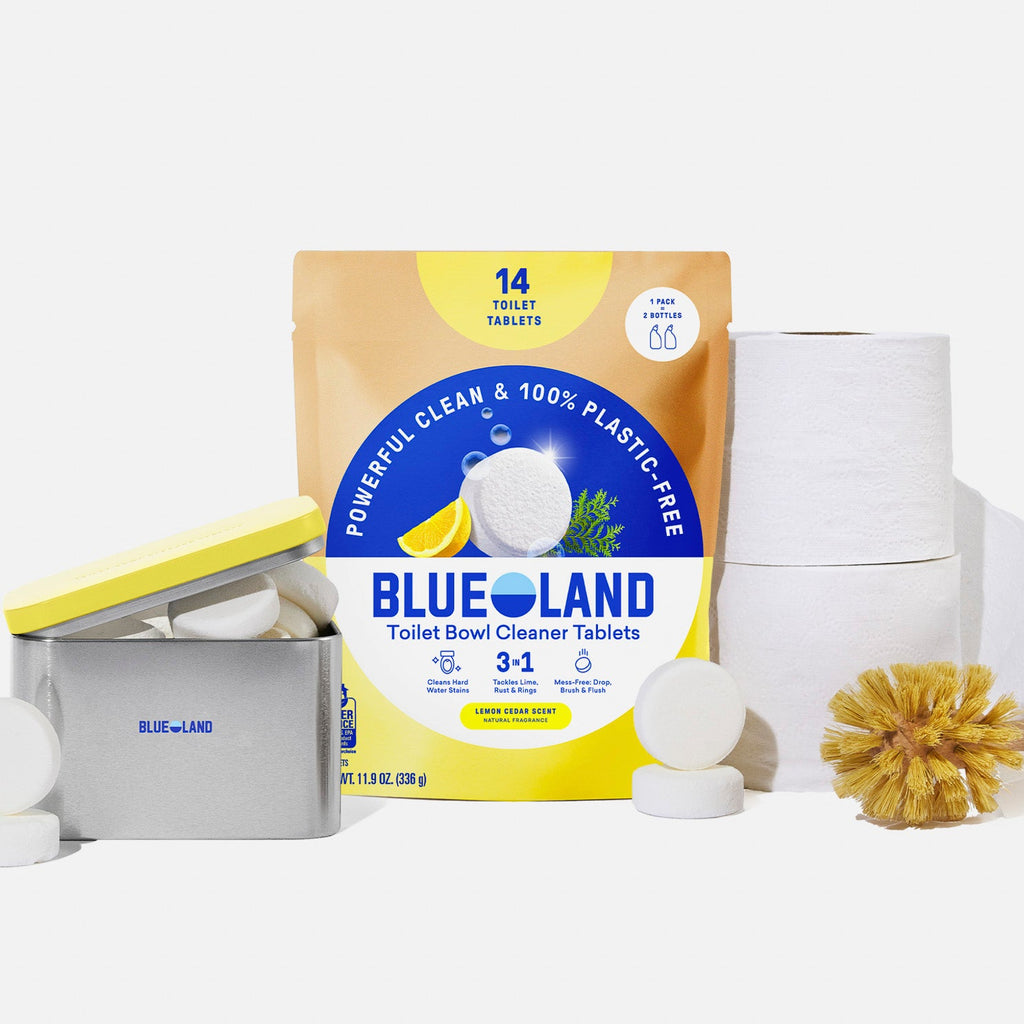 A package of Blueland toilet bowl cleaner tablets is shown with toilet paper rolls, a round scrub brush, a metal tin, and several white cleaning tablets in front of a white background.