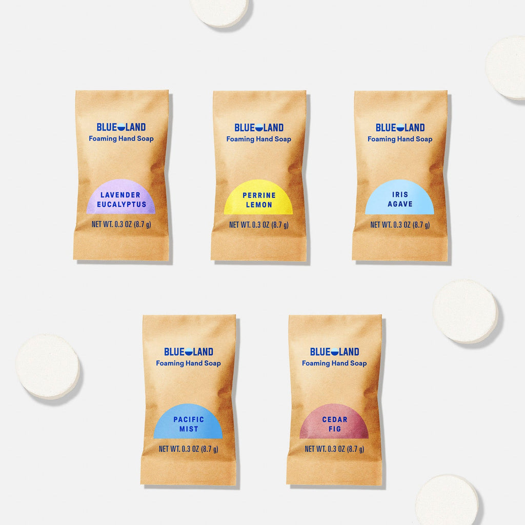 Five Blueland foaming hand soap refill packets in different scents—Lavender Eucalyptus, Perrine Lemon, Iris Agave, Pacific Mist, and Cedar Fig—are arranged on a light surface with round white soap tablets.