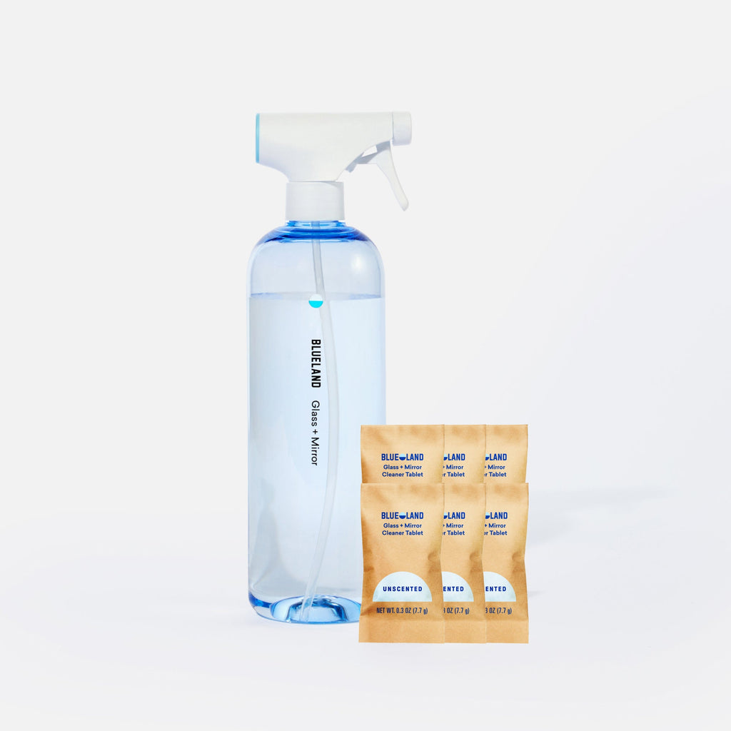 A clear spray bottle labeled Blueland Glass + Mirror filled with blue liquid stands next to four brown refill packets labeled Blueland Glass + Mirror Cleaner Tablet Unscented on a white background.