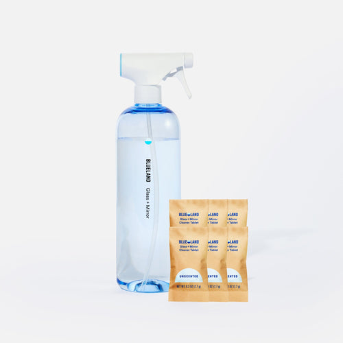 A clear spray bottle labeled Blueland Glass + Mirror filled with blue liquid stands next to four brown refill packets labeled Blueland Glass + Mirror Cleaner Tablet Unscented on a white background.