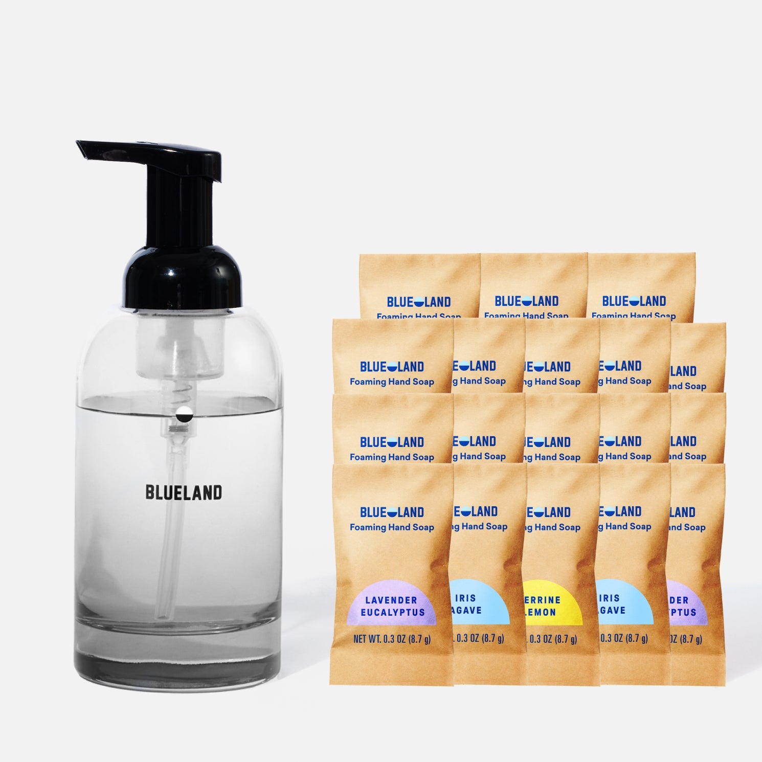A clear glass Blueland soap dispenser with a black pump is next to nine Blueland foaming hand soap refill packets in various scents, arranged in three rows, against a white background.
