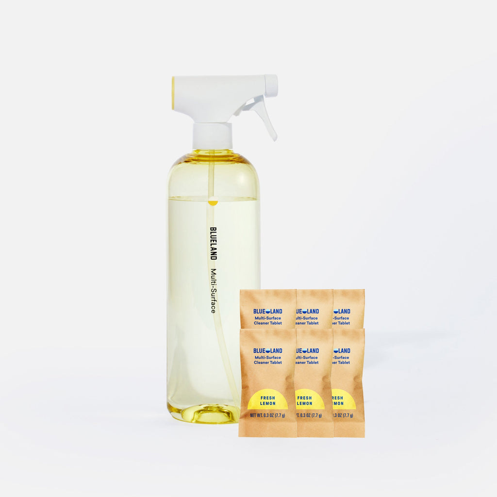 A clear spray bottle labeled Blueland Multi-Surface stands next to six small yellow and orange Blueland cleaning tablets in paper packets, all set against a white background.