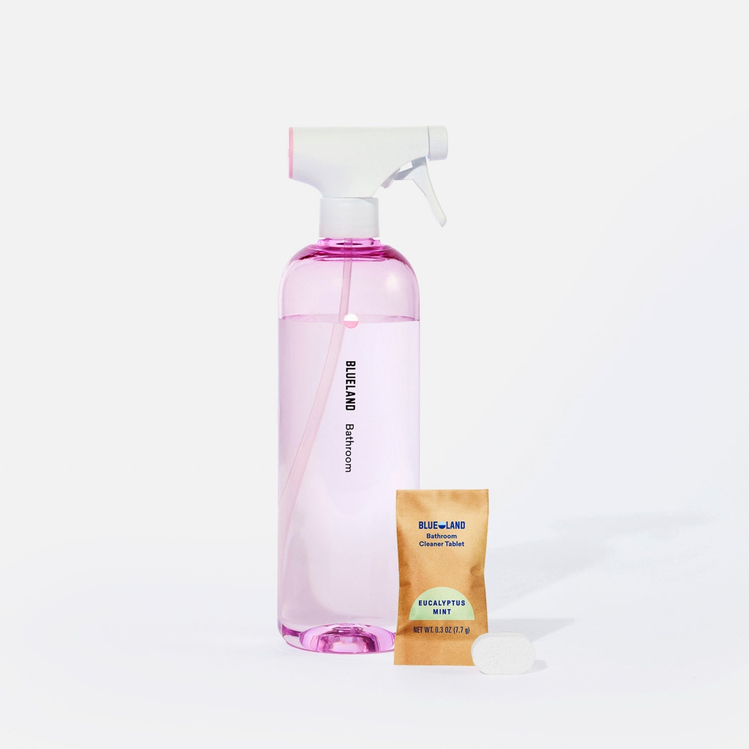 A clear spray bottle labeled “Blueland Bathroom” filled with pink liquid stands beside a small brown packet labeled Blueland Eucalyptus Mint Cleaner Tablet and a pink cleaning tablet.